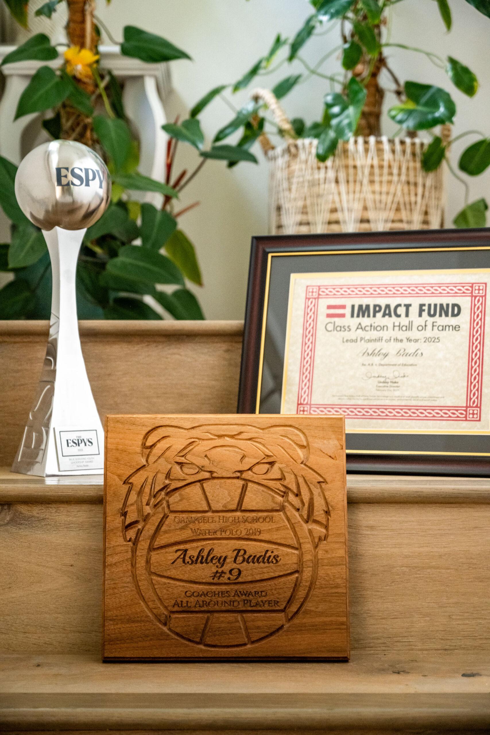 Photo of Ashley Badis's awards sitting on the steps in her family home with a 2019 James Campbell High School Coaches Award for All Around Player, her ESPYs award, and an Impact Fund Class Action Hall of Fame Lead Plaintiff of the Year: 2025 award.