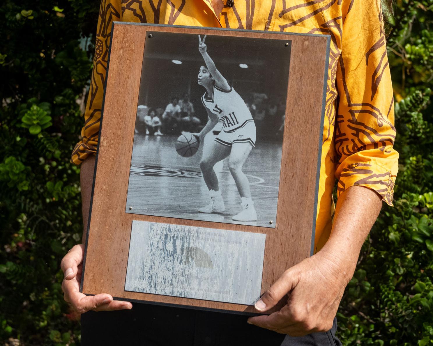 Portrait of Jill Nunokawa, a public defender and civil rights specialist at the University of Hawaii, holding a picture frame with a photo from her time as a Rainbow Wahine basketball player and early Title IX athlete.